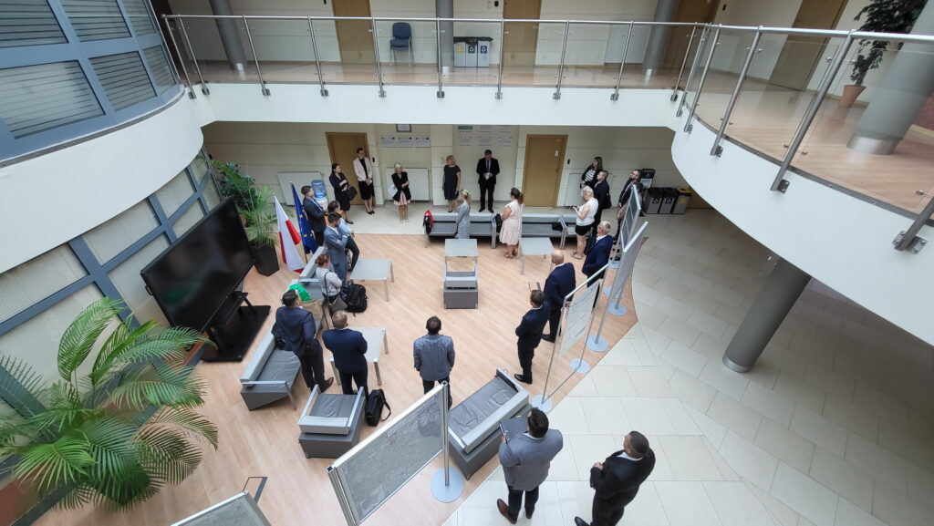 Zdjęcie przedstawia hol w siedzibie GUGiK przy ul. Olbrachta 94B z osobami przystępującymi do egzaminu słuchającymi wyjaśnień członków Komisji Egzaminacyjnej.