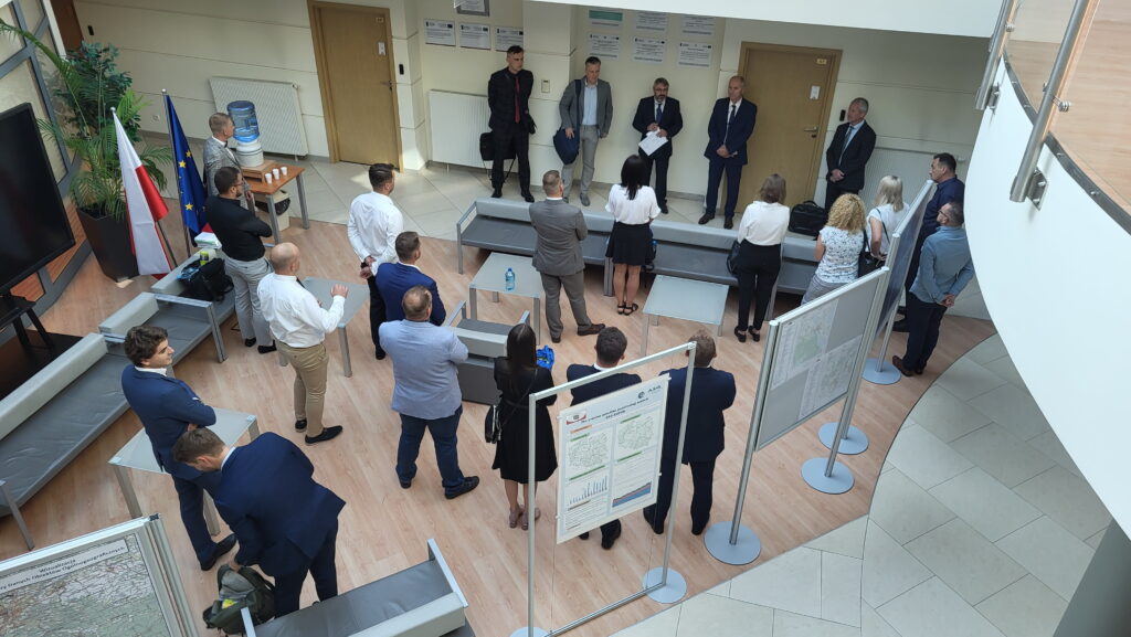 Zdjęcie przedstawia hol w siedzibie GUGiK przy ul. Olbrachta 94B z osobami przystępującymi do egzaminu słuchającymi wyjaśnień członków Komisji Egzaminacyjnej