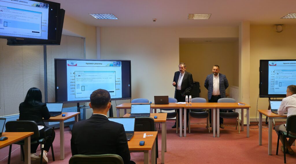 Fot. 2. Zdjęcie przedstawia salę egzaminacyjną z członkami Komisji egzaminacyjnej i osobami przystępującymi do egzaminu siedzącymi pojedynczo przed laptopami.