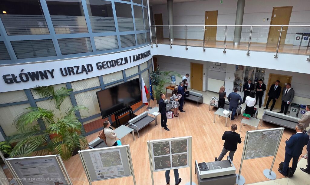 Zdjęcie przedstawia hol w siedzibie GUGiK przy ul. Olbrachta 94B z osobami przystępującymi do egzaminu słuchającymi wyjaśnień członków Komisji Egzaminacyjnej.