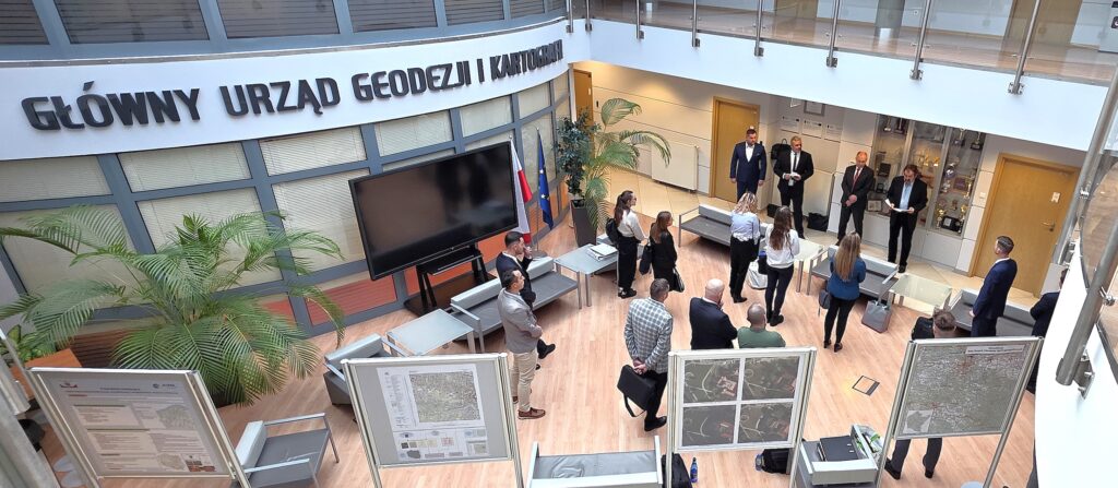 Zdjęcie przedstawia hol w siedzibie GUGiK przy ul. Olbrachta 94B z osobami przystępującymi do egzaminu słuchającymi wyjaśnień członków Komisji Egzaminacyjnej.