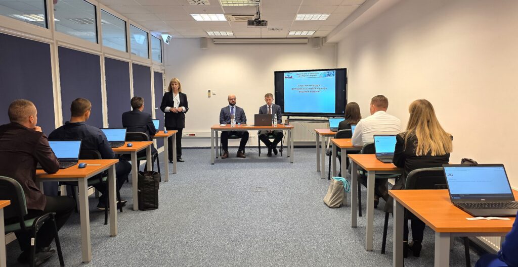 Zdjęcie przedstawia salę egzaminacyjną z członkami Komisji egzaminacyjnej i osobami przystępującymi do egzaminu siedzącymi pojedynczo przed laptopami.