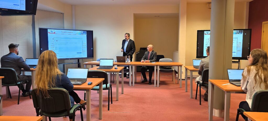 Zdjęcie przedstawia salę egzaminacyjną z członkami Komisji egzaminacyjnej i osobami przystępującymi do egzaminu siedzącymi pojedynczo przed laptopami.