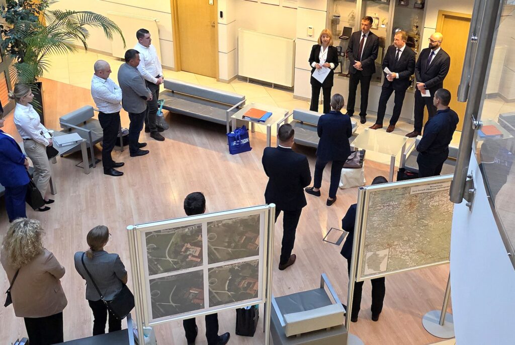 Zdjęcie przedstawia hol w siedzibie GUGiK przy ul. Olbrachta 94B z osobami przystępującymi do egzaminu słuchającymi wyjaśnień członków Komisji Egzaminacyjnej.