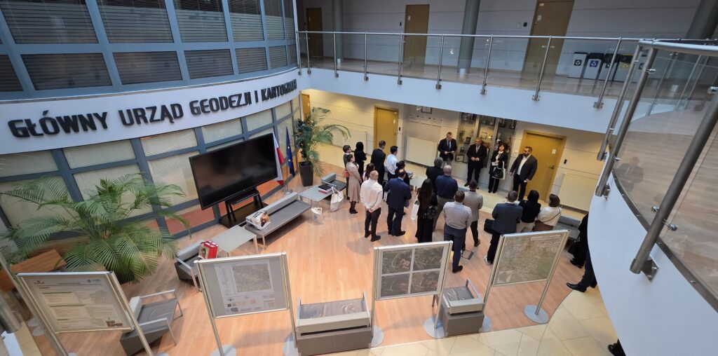 Zdjęcie przedstawia hol w siedzibie GUGiK przy ul. Olbrachta 94B z osobami przystępującymi do egzaminu słuchającymi wyjaśnień członków Komisji Egzaminacyjnej.