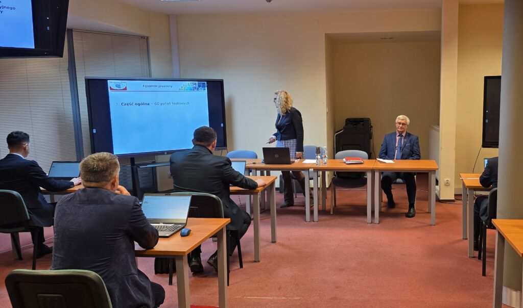 Zdjęcie przedstawia salę egzaminacyjną z członkami Komisji egzaminacyjnej i osobami przystępującymi do egzaminu siedzącymi pojedynczo przed laptopami.