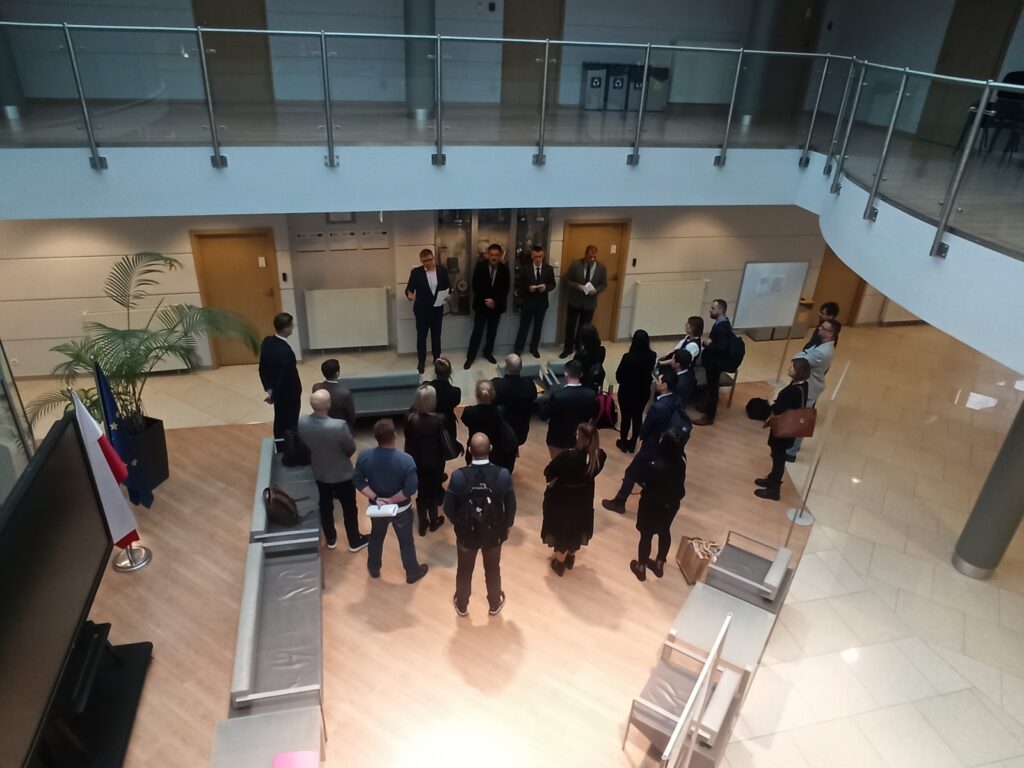 Zdjęcie przedstawia hol w siedzibie GUGiK przy ul. Olbrachta 94B z osobami przystępującymi do egzaminu słuchającymi wyjaśnień członków Komisji Egzaminacyjnej.