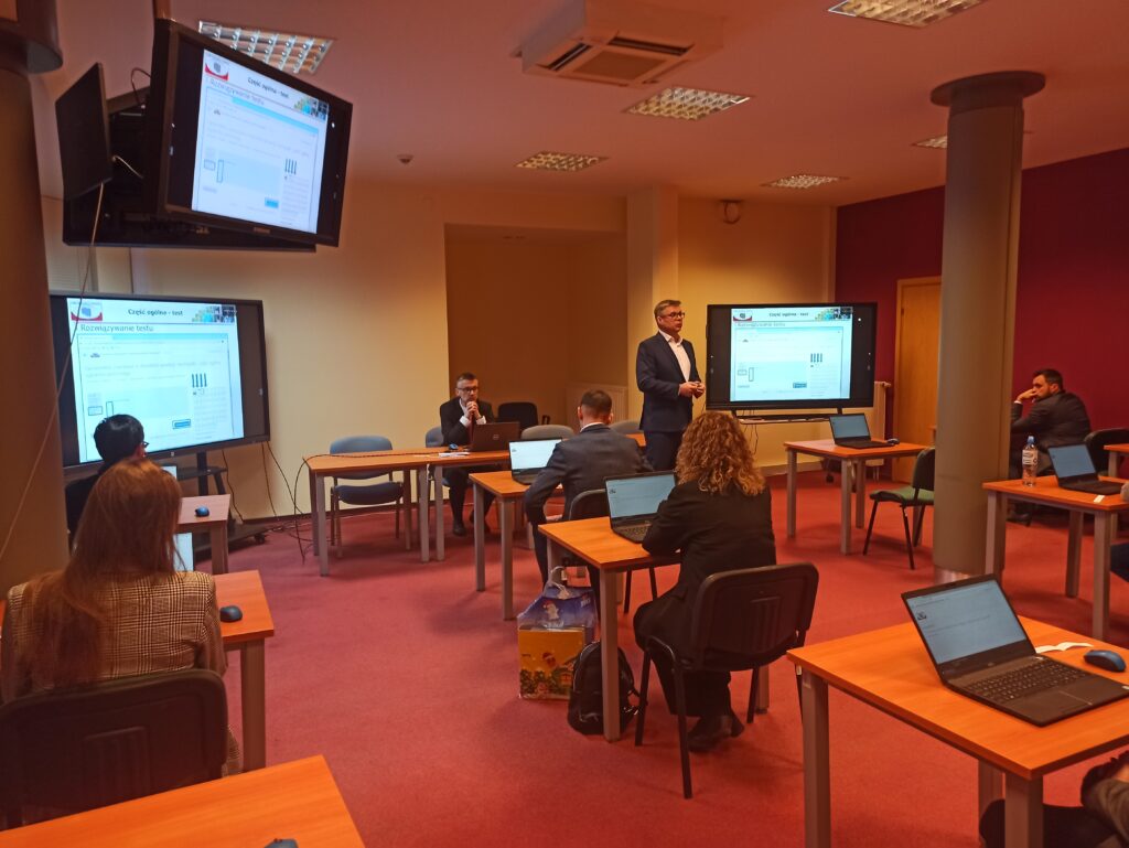 Zdjęcie przedstawia salę egzaminacyjną z członkami Komisji egzaminacyjnej i osobami przystępującymi do egzaminu siedzącymi pojedynczo przed laptopami.