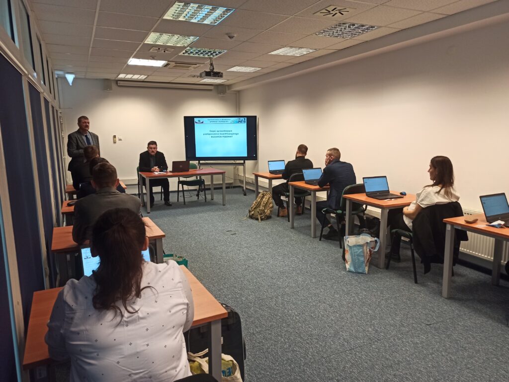 Zdjęcie przedstawia salę egzaminacyjną z członkami Komisji egzaminacyjnej i osobami przystępującymi do egzaminu siedzącymi pojedynczo przed laptopami.