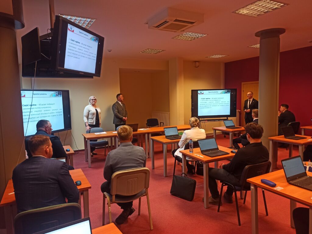 Zdjęcie przedstawia salę egzaminacyjną z członkami Komisji egzaminacyjnej i osobami przystępującymi do egzaminu siedzącymi pojedynczo przed laptopami.