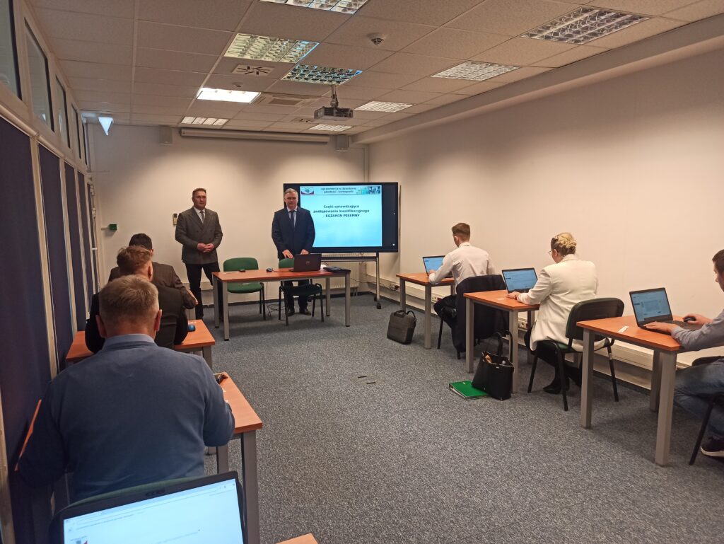 Zdjęcie przedstawia salę egzaminacyjną z członkami Komisji egzaminacyjnej i osobami przystępującymi do egzaminu siedzącymi pojedynczo przed laptopami.