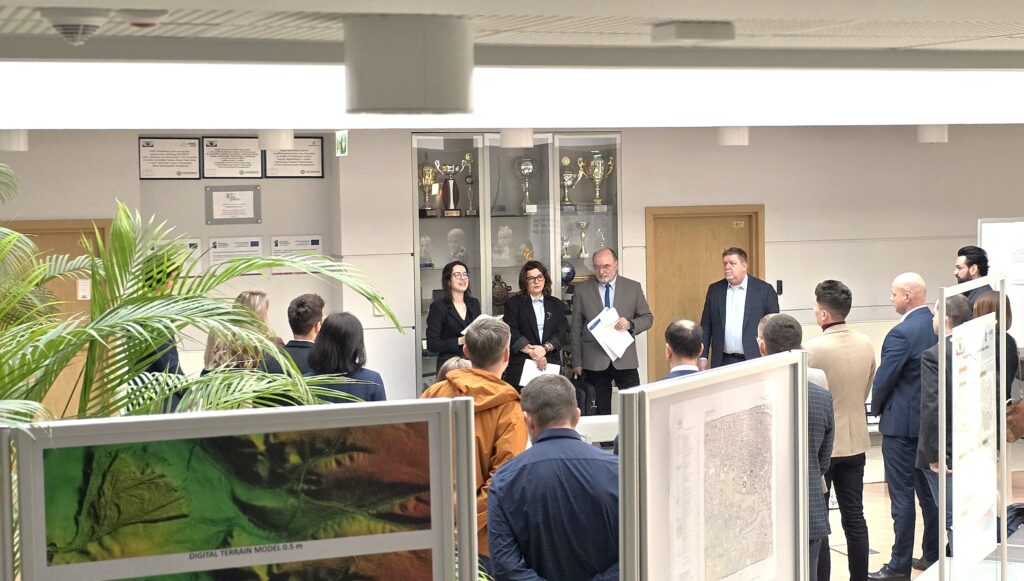 Zdjęcie przedstawia hol w siedzibie GUGiK przy ul. Olbrachta 94B z osobami przystępującymi do egzaminu słuchającymi wyjaśnień członków Komisji Egzaminacyjnej.