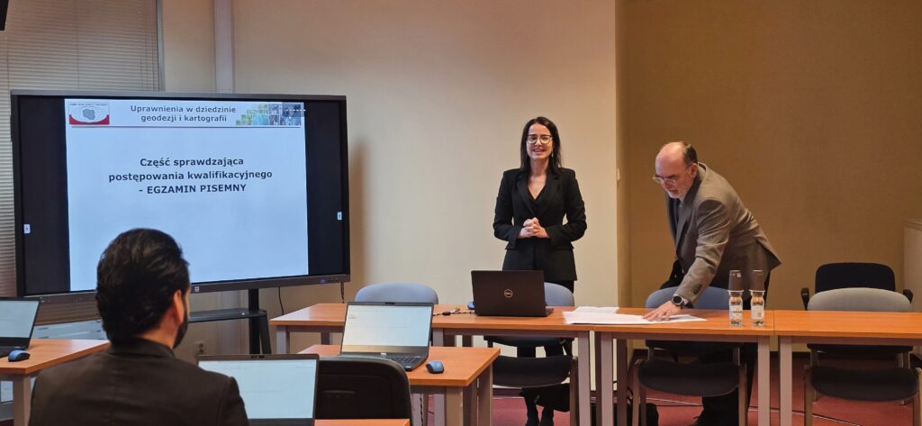 Zdjęcie przedstawia salę egzaminacyjną z członkami Komisji egzaminacyjnej i osobami przystępującymi do egzaminu siedzącymi pojedynczo przed laptopami.