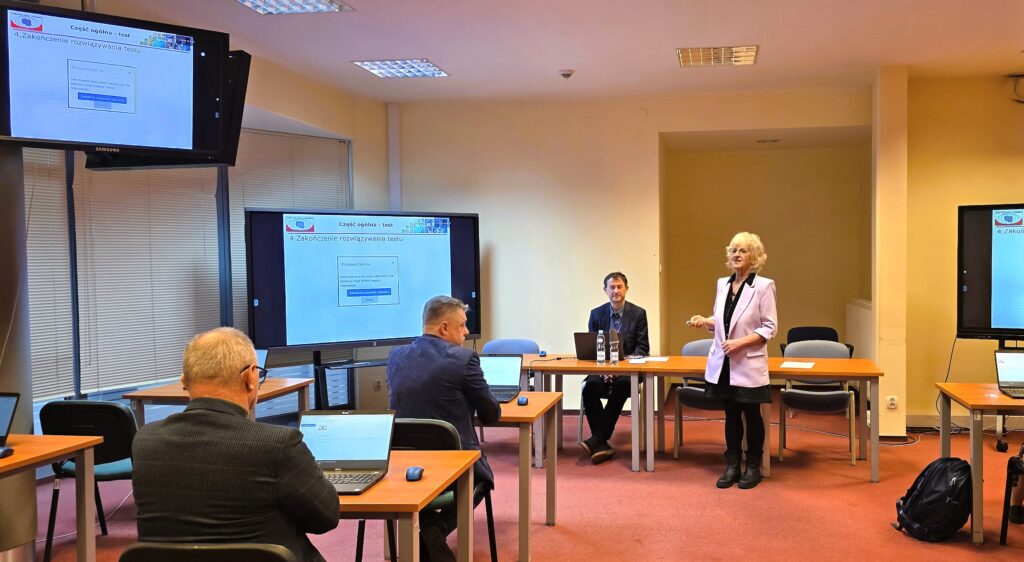 Zdjęcie przedstawia salę egzaminacyjną z członkami Komisji egzaminacyjnej i osobami przystępującymi do egzaminu siedzącymi pojedynczo przed laptopami.