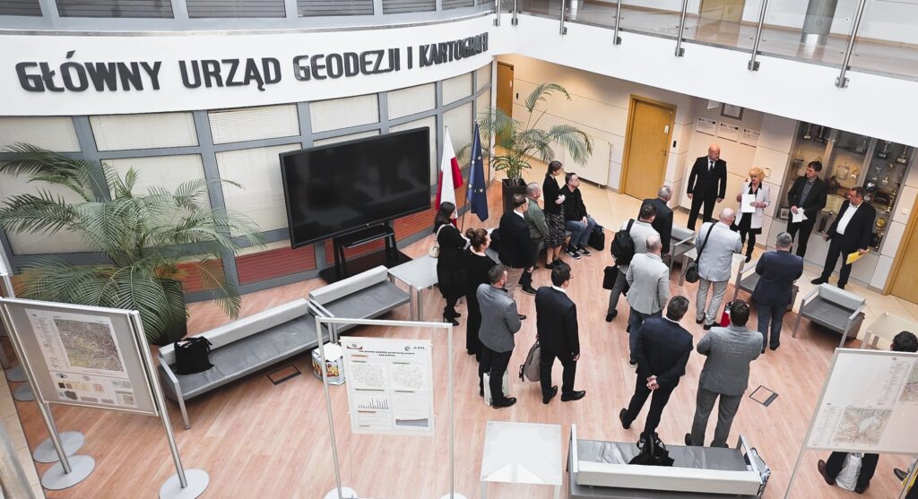 Zdjęcie przedstawia hol w siedzibie GUGiK przy ul. Olbrachta 94B z osobami przystępującymi do egzaminu słuchającymi wyjaśnień członków Komisji Egzaminacyjnej.