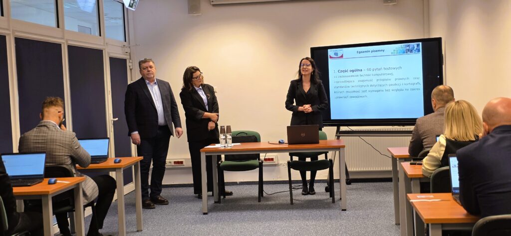 Zdjęcie przedstawia salę egzaminacyjną z członkami Komisji egzaminacyjnej i osobami przystępującymi do egzaminu siedzącymi pojedynczo przed laptopami.