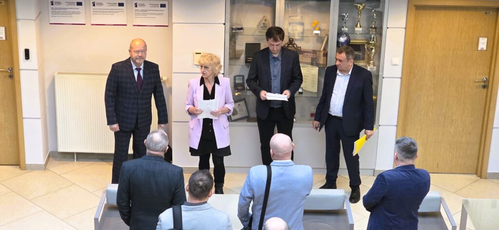 Zdjęcie przedstawia hol w siedzibie GUGiK przy ul. Olbrachta 94B z osobami przystępującymi do egzaminu słuchającymi wyjaśnień członków Komisji Egzaminacyjnej.