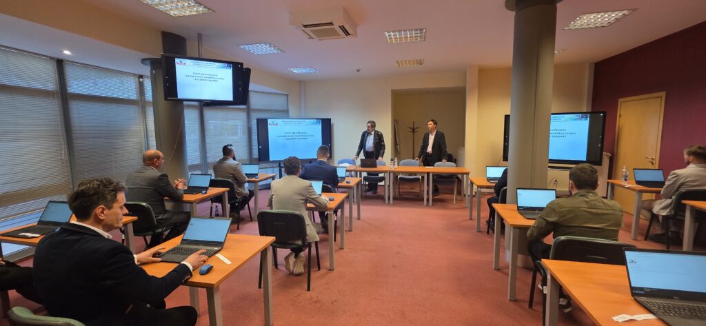 Fot. 2. Zdjęcie przedstawia salę egzaminacyjną z członkami Komisji egzaminacyjnej i osobami przystępującymi do egzaminu siedzącymi pojedynczo przed laptopami.