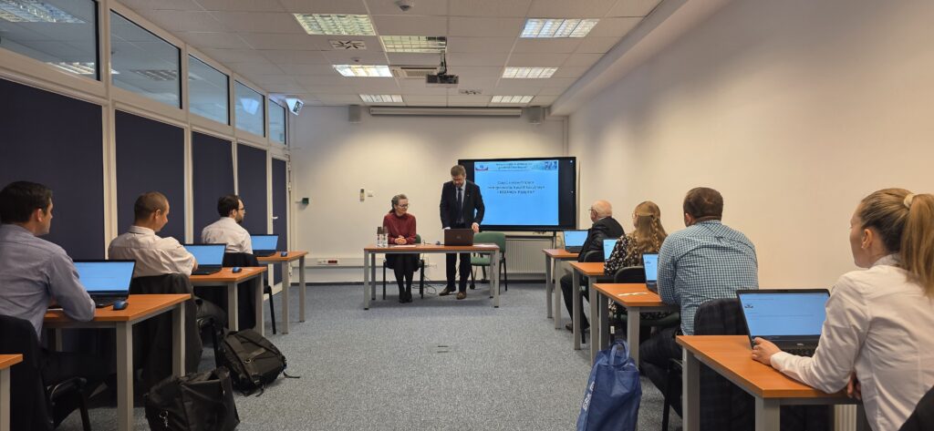 Fot. 3. Zdjęcie przedstawia salę egzaminacyjną z członkami Komisji egzaminacyjnej i osobami przystępującymi do egzaminu siedzącymi pojedynczo przed laptopami.