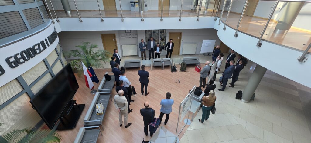 Fot. 4. Zdjęcie przedstawia hol w siedzibie GUGiK przy ul. Olbrachta 94B z osobami przystępującymi do egzaminu słuchającymi wyjaśnień członków Komisji Egzaminacyjnej.