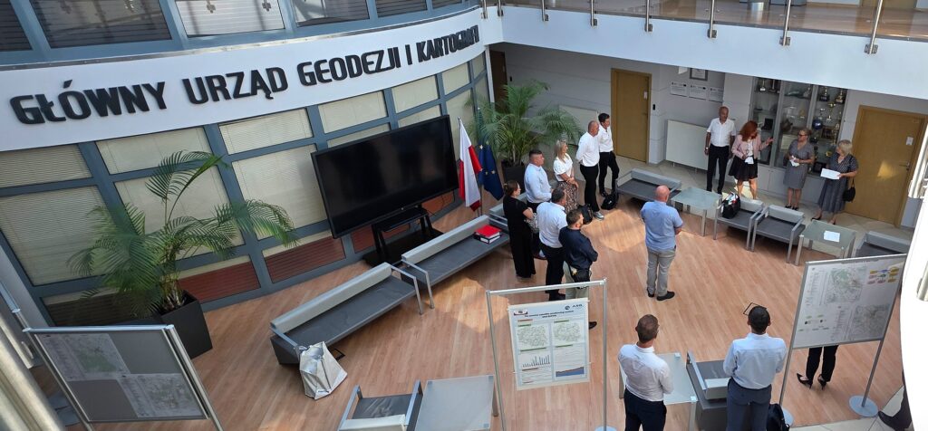 Zdjęcie przedstawia hol w siedzibie GUGiK przy ul. Olbrachta 94B z osobami przystępującymi do egzaminu słuchającymi wyjaśnień członków Komisji Egzaminacyjnej.