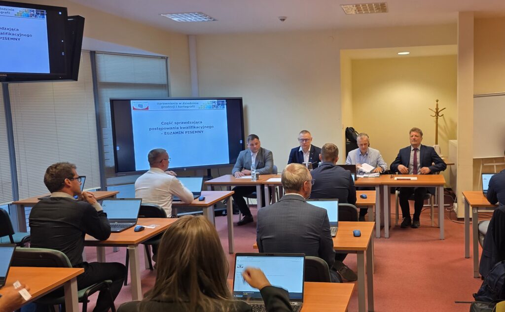 Zdjęcie przedstawia salę egzaminacyjną z członkami Komisji egzaminacyjnej i osobami przystępującymi do egzaminu siedzącymi pojedynczo przed laptopami.