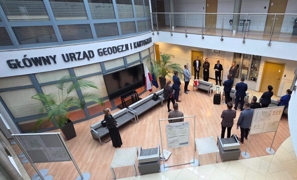 Zdjęcie przedstawia hol w siedzibie GUGiK przy ul. Olbrachta 94B z osobami przystępującymi do egzaminu słuchającymi wyjaśnień członków Komisji Egzaminacyjnej.