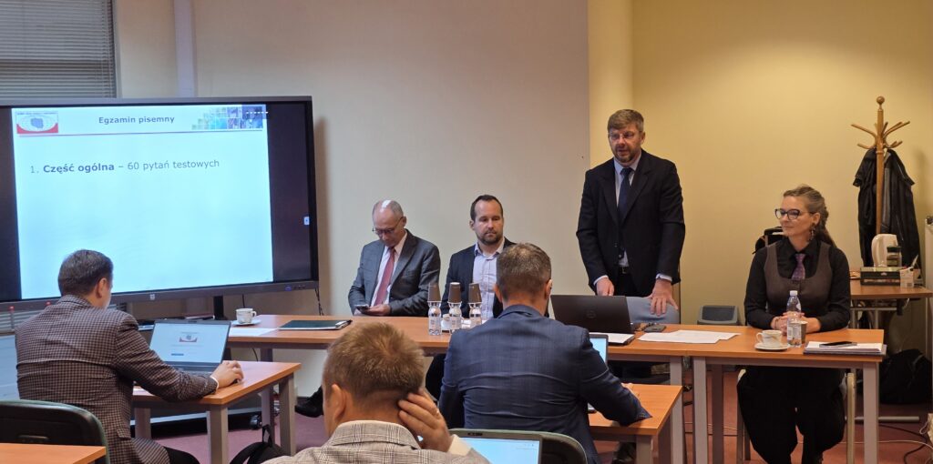 Zdjęcie przedstawia salę egzaminacyjną z członkami Komisji egzaminacyjnej i osobami przystępującymi do egzaminu siedzącymi pojedynczo przed laptopami.