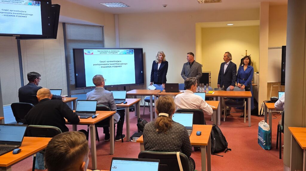 Zdjęcie przedstawia salę egzaminacyjną z członkami Komisji egzaminacyjnej i osobami przystępującymi do egzaminu siedzącymi pojedynczo przed laptopami.