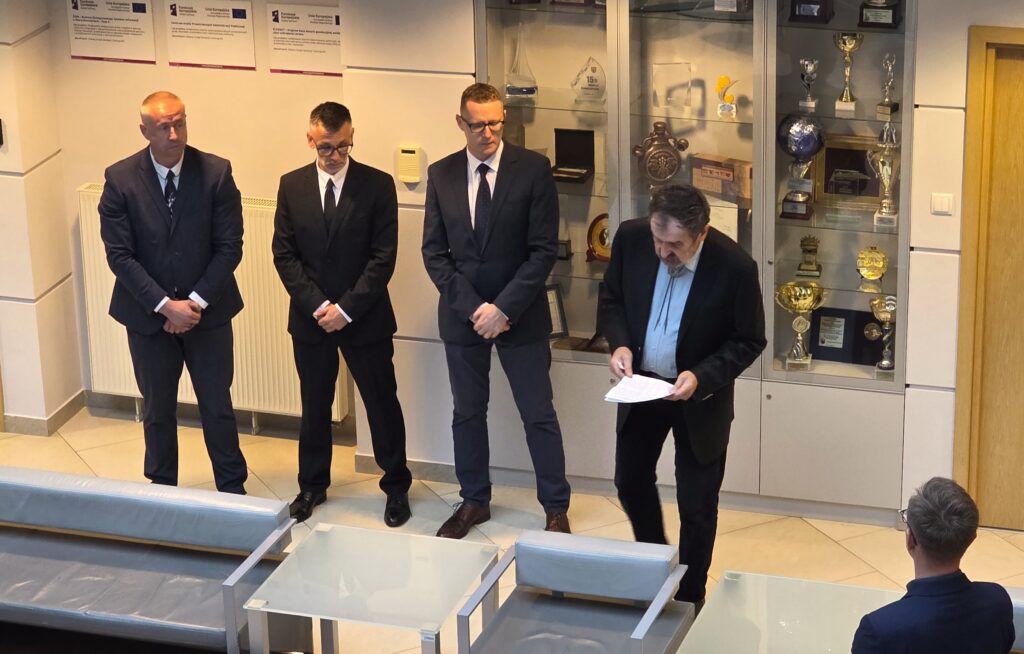 Zdjęcie przedstawia hol w siedzibie GUGiK przy ul. Olbrachta 94B z osobami przystępującymi do egzaminu słuchającymi wyjaśnień członków Komisji Egzaminacyjnej.