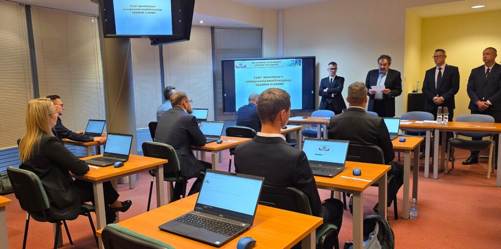 Zdjęcie przedstawia salę egzaminacyjną z członkami Komisji egzaminacyjnej i osobami przystępującymi do egzaminu siedzącymi pojedynczo przed laptopami.