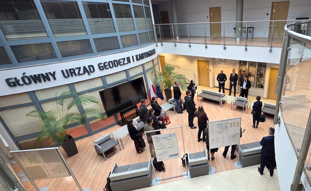Zdjęcie przedstawia hol w siedzibie GUGiK przy ul. Olbrachta 94B z osobami przystępującymi do egzaminu słuchającymi wyjaśnień członków Komisji Egzaminacyjnej.